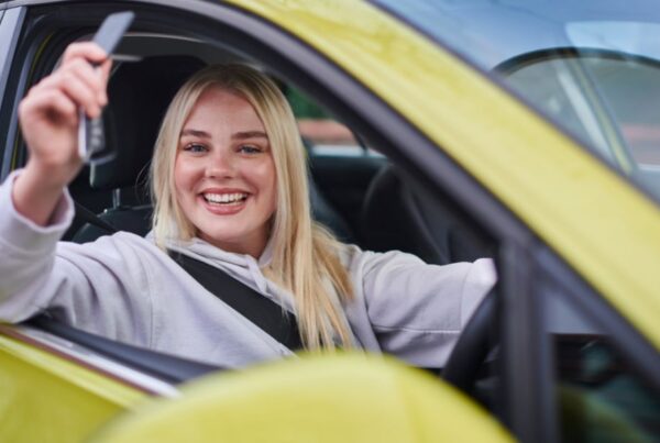 Smiling blonde woman sitting in the driver's seat of a yellow car, holding up a car key through the open window