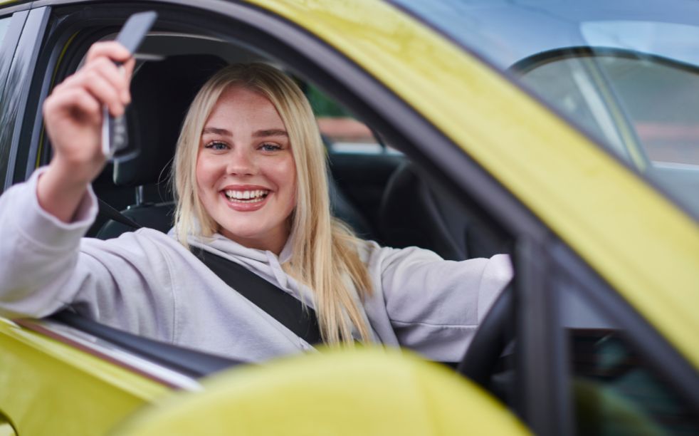 Smiling blonde woman sitting in the driver's seat of a yellow car, holding up a car key through the open window