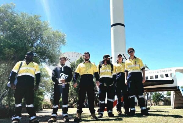 A group of six individuals standing side-by-side outdoors on a grassy lawn. Most of them are dressed in high-visibility work uniforms consisting of yellow and dark blue/black sections.