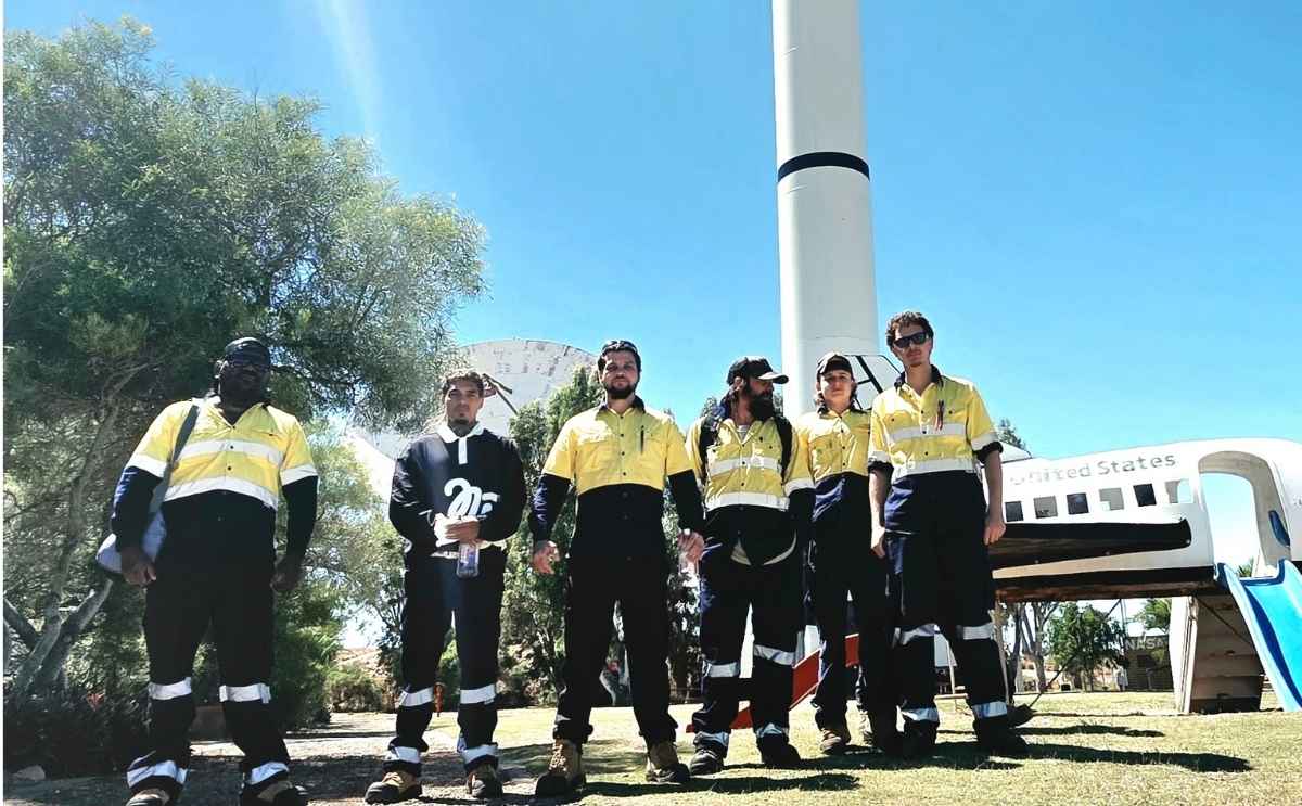 A group of six individuals standing side-by-side outdoors on a grassy lawn. Most of them are dressed in high-visibility work uniforms consisting of yellow and dark blue/black sections.